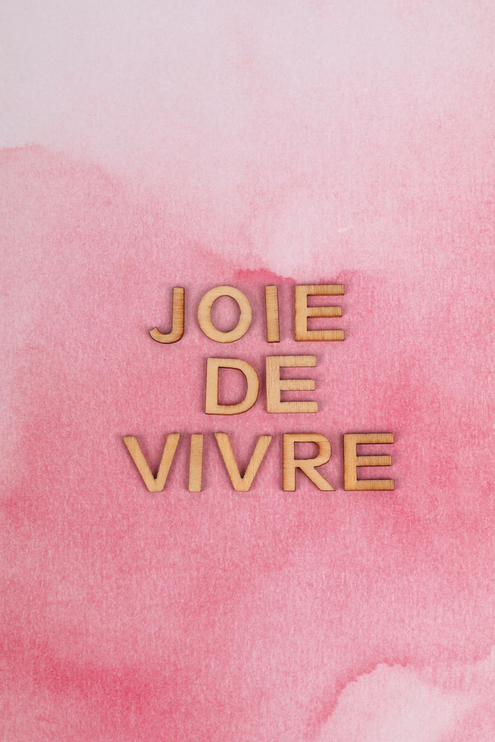 Wooden letters spelling 'Joie De Vivre' on a pink watercolor background for creative decoration.